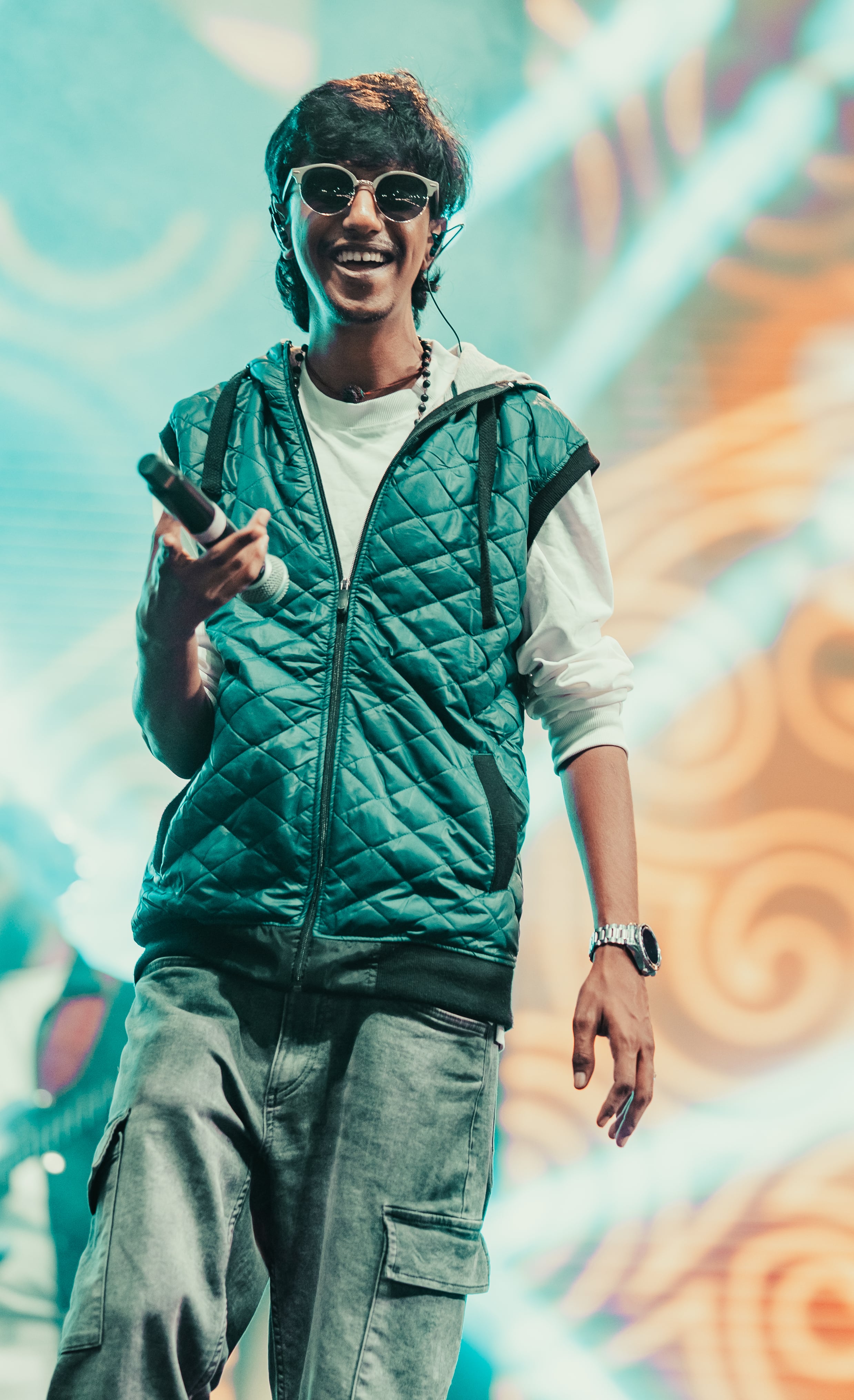 Sanikelama grinning on stage with a mic, in a teal quilted vest and white sunglasses