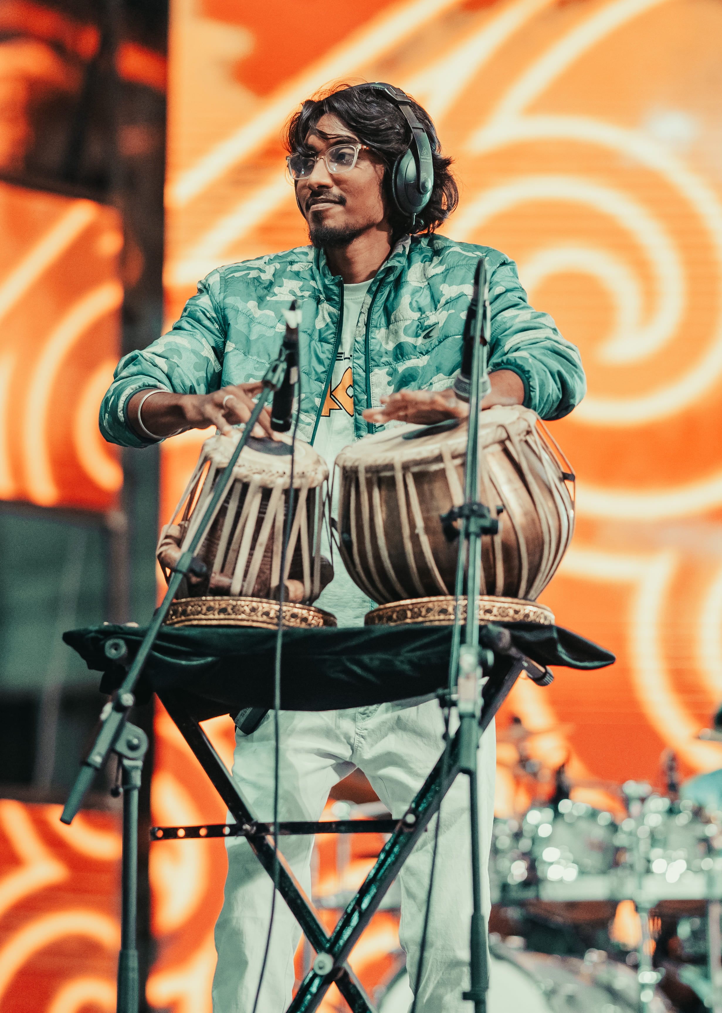 Vasanth in headphones and a teal camo jacket playing tabla on stage